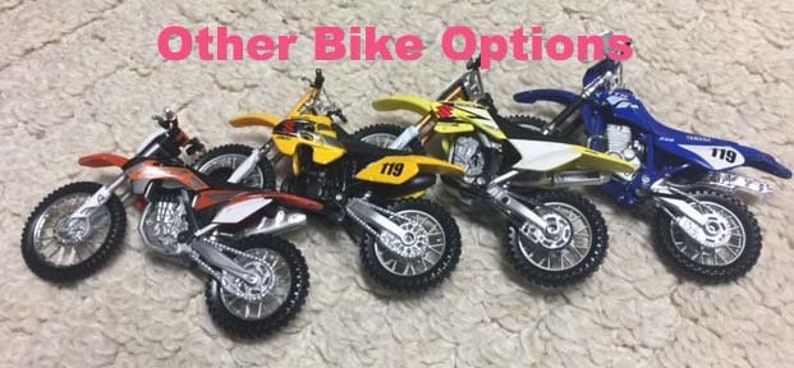 May include: Four toy dirt bikes in different colors, including orange, yellow, and blue. Each bike has a number "119" printed on it. The bikes are arranged in a row on a brown surface. The text "Other Bike Options" is written in pink above the bikes.