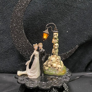 May include: A black crescent moon with glitter, a bride and groom figurine, and a lamppost with a stack of skulls. The base is black with a decorative design.
