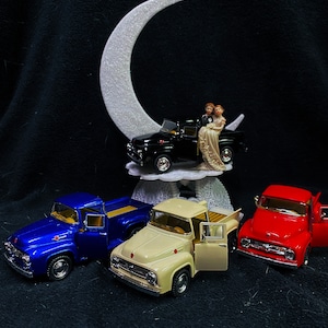 May include: Three toy pickup trucks, one blue, one cream, and one red, are arranged on a black surface. A white crescent moon sits above the trucks, with a miniature bride and groom figurine in a black pickup truck on top of the moon.