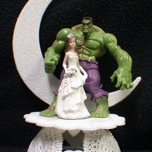 May include: A white cake topper featuring a green Hulk figurine holding a bride figurine wearing a white dress. The couple are standing on a white crescent moon base with a white lace base.