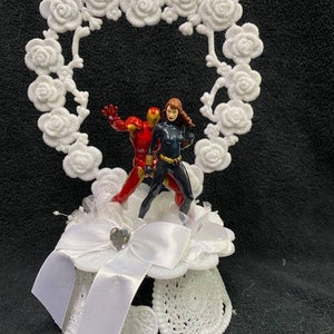 May include: White cake topper featuring a heart-shaped frame of white roses with a red and black superhero couple standing inside. The base of the topper is white with a white bow and a rhinestone.