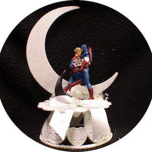 May include: A white and silver crescent moon cake topper with two figures in superhero costumes kissing. The figures are standing on the moon and are surrounded by white lace.