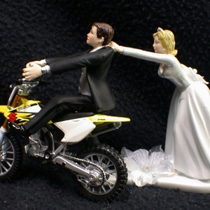 May include: A yellow and white Suzuki dirt bike wedding cake topper with a groom figurine riding the bike and a bride figurine reaching out to him.
