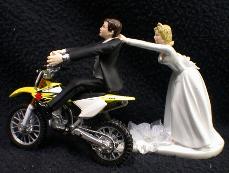 May include: A yellow and white Suzuki dirt bike wedding cake topper with a groom figurine riding the bike and a bride figurine reaching out to him.