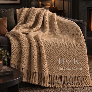 May include: A beige knit throw blanket with a textured popcorn stitch pattern and long fringe along the bottom edge. The blanket features the white embroidered text H ♡ K and the words Our Cozy Corner centred on the lower right side.