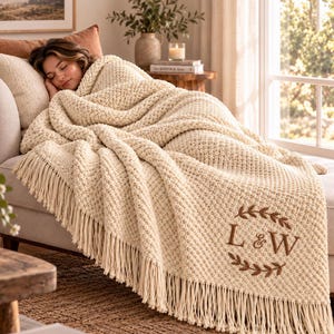 May include: A cream-coloured chunky knit throw blanket featuring a textured popcorn stitch pattern and long fringe along the edges. The blanket is embroidered with a brown laurel wreath design surrounding the letters L & W in the bottom corner.