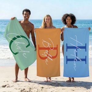 May include: Three beach towels in shades of green, orange, and blue are held up on a sandy beach. Each towel features a large initial and a name in a contrasting color. The towels are rectangular and appear to be made of a soft fabric.