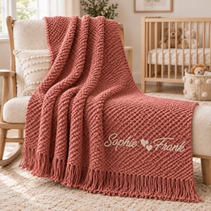 Può includere: Una coperta in maglia grossa color rosa polvere con un motivo a punto popcorn testurizzato e lunghe frange sul bordo inferiore. La coperta è ricamata con il testo in corsivo bianco "Sophie" seguito da due piccoli cuori e "Frank".