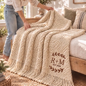 May include: A cream-coloured chunky knit throw blanket featuring a fringe edge and an embroidered brown design with the letters R+M and the text EST. 2026 enclosed within a decorative leaf wreath motif on the lower right corner of the fabric.