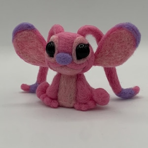 Puede incluir: Un personaje de fieltro rosa hecho a mano con grandes ojos negros, detalles morados y orejas grandes y ca&iacute;das. El personaje est&aacute; sentado con los brazos y las piernas extendidos. El fieltro es un material suave y texturizado. El dise&ntilde;o es caprichoso y lindo.