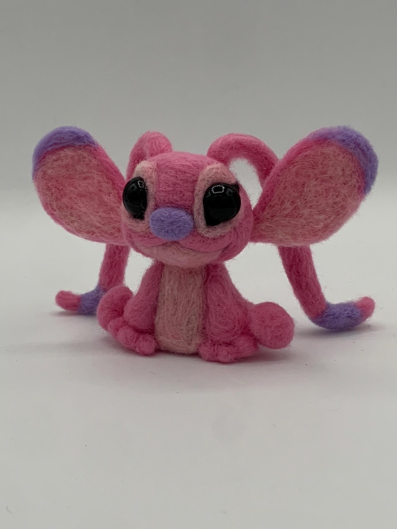 Puede incluir: Un personaje de fieltro rosa hecho a mano con grandes ojos negros, detalles morados y orejas grandes y ca&iacute;das. El personaje est&aacute; sentado con los brazos y las piernas extendidos. El fieltro es un material suave y texturizado. El dise&ntilde;o es caprichoso y lindo.