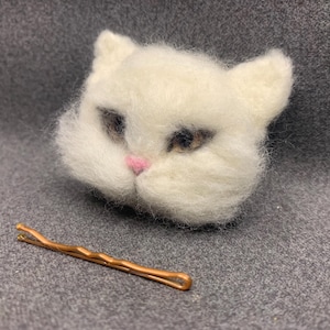 May include: A white felted cat head with brown eyes and a pink nose. A copper-colored bobby pin rests below the cat. The cat's fur is fluffy and detailed, creating a whimsical accessory.