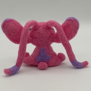 Puede incluir: Una criatura de fieltro rosa hecha a mano con orejas y extremidades largas y curvas. Las puntas de las orejas y las extremidades son moradas. La criatura tiene un parche morado en la espalda y una peque&ntilde;a cola redonda. El juguete est&aacute; sobre un fondo blanco liso.