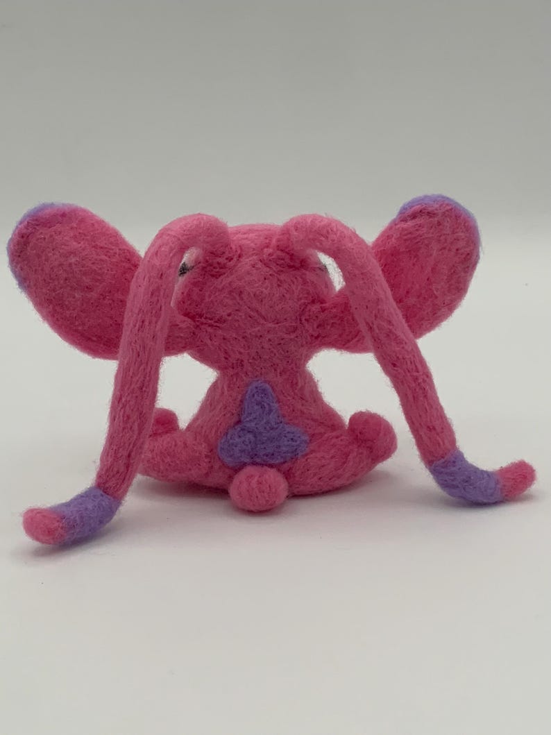 Puede incluir: Una criatura de fieltro rosa hecha a mano con orejas y extremidades largas y curvas. Las puntas de las orejas y las extremidades son moradas. La criatura tiene un parche morado en la espalda y una peque&ntilde;a cola redonda. El juguete est&aacute; sobre un fondo blanco liso.
