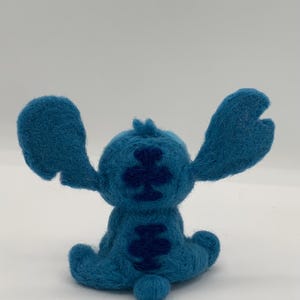 Puede incluir: Figura de Stitch de fieltro azul hecha a mano, con orejas grandes y una cola peque&ntilde;a. La espalda del personaje tiene un patr&oacute;n azul m&aacute;s oscuro. La figura est&aacute; sobre una superficie blanca.