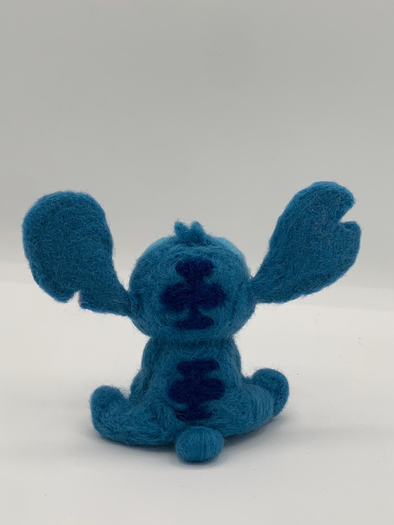 Puede incluir: Figura de Stitch de fieltro azul hecha a mano, con orejas grandes y una cola peque&ntilde;a. La espalda del personaje tiene un patr&oacute;n azul m&aacute;s oscuro. La figura est&aacute; sobre una superficie blanca.
