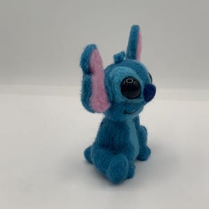 Puede incluir: Figura de Stitch de fieltro azul con grandes ojos negros, orejas internas rosas y una peque&ntilde;a nariz azul. El personaje hecho a mano est&aacute; sentado, mostrando sus rasgos distintivos y su expresi&oacute;n juguetona. Fondo blanco.