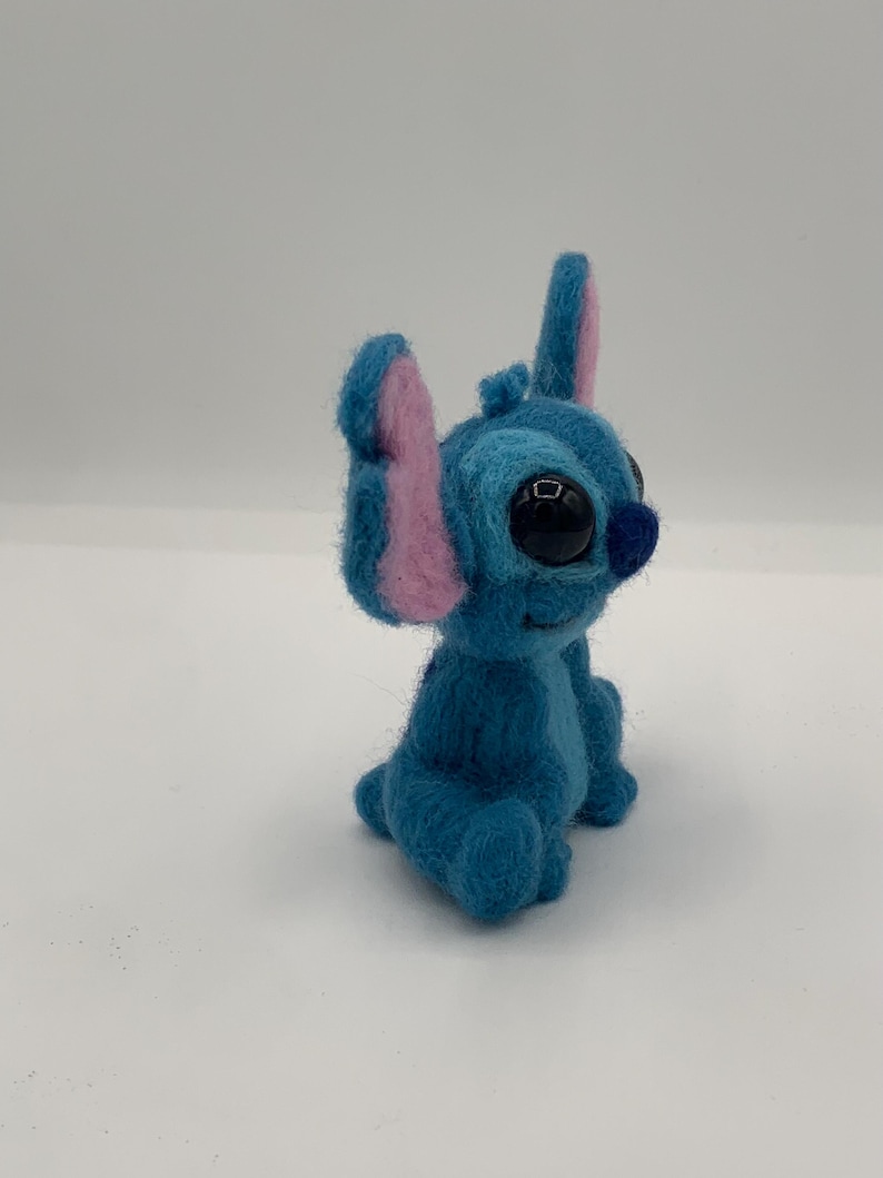 Puede incluir: Figura de Stitch de fieltro azul con grandes ojos negros, orejas internas rosas y una peque&ntilde;a nariz azul. El personaje hecho a mano est&aacute; sentado, mostrando sus rasgos distintivos y su expresi&oacute;n juguetona. Fondo blanco.