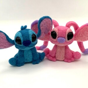 Puede incluir: Dos figuras de fieltro hechas a mano de personajes de dibujos animados. Una es azul con orejas grandes y la otra es rosa con orejas grandes. Ambos tienen ojos negros grandes y est&aacute;n sentados. Las figuras est&aacute;n sobre un fondo blanco.