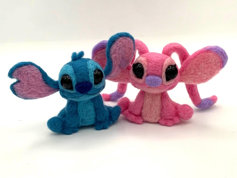 Puede incluir: Dos figuras de fieltro hechas a mano de personajes de dibujos animados. Una es azul con orejas grandes y la otra es rosa con orejas grandes. Ambos tienen ojos negros grandes y est&aacute;n sentados. Las figuras est&aacute;n sobre un fondo blanco.