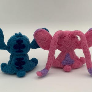 Puede incluir: Dos figuras de personajes de fieltro, una turquesa y otra rosa, est&aacute;n una al lado de la otra. La figura turquesa tiene orejas grandes y marcas azul oscuro. La figura rosa tiene orejas largas y detalles morados.