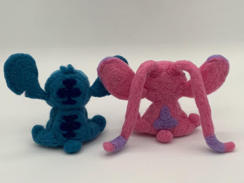 Puede incluir: Dos figuras de personajes de fieltro, una turquesa y otra rosa, est&aacute;n una al lado de la otra. La figura turquesa tiene orejas grandes y marcas azul oscuro. La figura rosa tiene orejas largas y detalles morados.