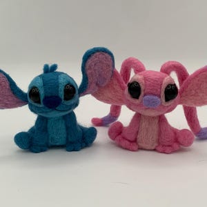 Puede incluir: Dos figuras de personajes de fieltro, una azul con orejas rosas y la otra rosa con detalles morados. Las figuras tienen grandes ojos negros y est&aacute;n sentadas. La figura azul est&aacute; a la izquierda y la rosa a la derecha.