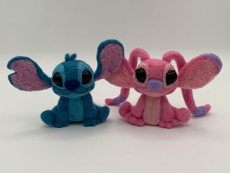 Puede incluir: Dos figuras de personajes de fieltro, una azul con orejas rosas y la otra rosa con detalles morados. Las figuras tienen grandes ojos negros y est&aacute;n sentadas. La figura azul est&aacute; a la izquierda y la rosa a la derecha.