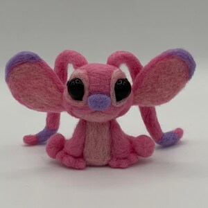 Puede incluir: Una criatura de fieltro rosa hecha a mano con grandes ojos negros, una nariz morada y orejas y extremidades con puntas moradas. El peluche est&aacute; sentado, mostrando sus detalles y su textura suave. El dise&ntilde;o es caprichoso y encantador.