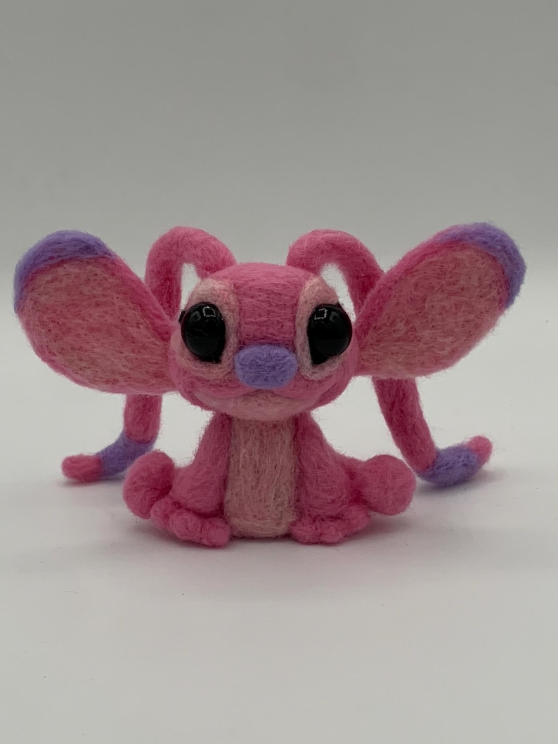 Puede incluir: Una criatura de fieltro rosa hecha a mano con grandes ojos negros, una nariz morada y orejas y extremidades con puntas moradas. El peluche est&aacute; sentado, mostrando sus detalles y su textura suave. El dise&ntilde;o es caprichoso y encantador.