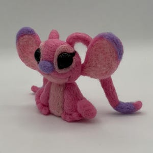 Puede incluir: Figura de fieltro rosa hecha a mano de un personaje de dibujos animados con orejas grandes, ojos negros y nariz morada. La figura est&aacute; sentada con los brazos y las piernas extendidos. Las orejas y los pies tienen detalles morados.