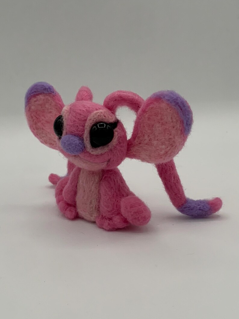 Puede incluir: Figura de fieltro rosa hecha a mano de un personaje de dibujos animados con orejas grandes, ojos negros y nariz morada. La figura est&aacute; sentada con los brazos y las piernas extendidos. Las orejas y los pies tienen detalles morados.