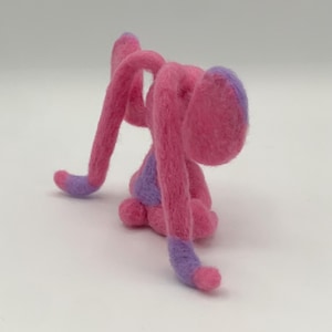 Puede incluir: Una escultura de fieltro hecha a mano de una criatura caprichosa en tonos de rosa y morado. La criatura tiene extremidades largas y curvas y una cabeza redondeada, con detalles morados en las puntas.