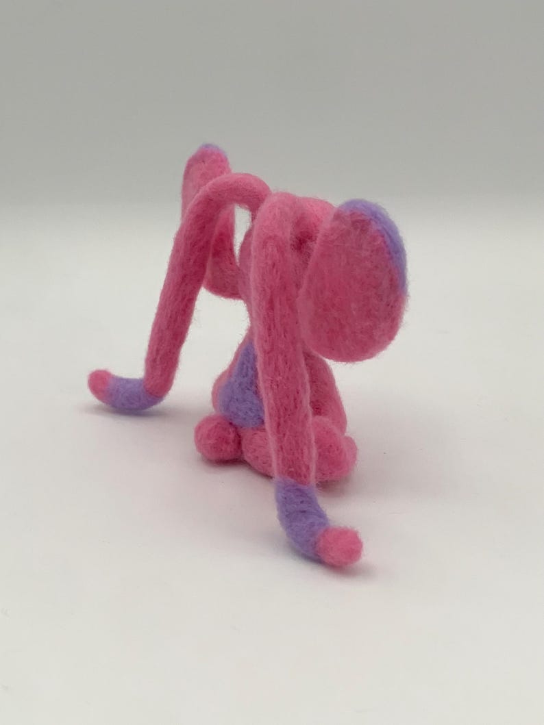 Puede incluir: Una escultura de fieltro hecha a mano de una criatura caprichosa en tonos de rosa y morado. La criatura tiene extremidades largas y curvas y una cabeza redondeada, con detalles morados en las puntas.