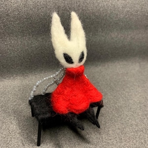 Peut inclure: Figurine artisanale en feutre représentant un personnage avec une tête blanche, des yeux noirs et une robe rouge. La figurine est assise sur un petit banc noir à quatre pieds. Une chaîne de perles est attachée.