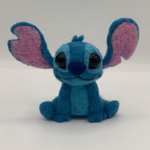 Puede incluir: Figura de Stitch hecha a mano en fieltro en tonos azules y rosas. El personaje tiene ojos negros grandes y expresivos, una nariz azul y orejas grandes y erguidas con secciones internas rosas. La figura est&aacute; sentada con los brazos y las piernas extendidos.