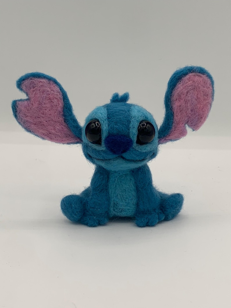 Puede incluir: Figura de Stitch hecha a mano en fieltro en tonos azules y rosas. El personaje tiene ojos negros grandes y expresivos, una nariz azul y orejas grandes y erguidas con secciones internas rosas. La figura est&aacute; sentada con los brazos y las piernas extendidos.