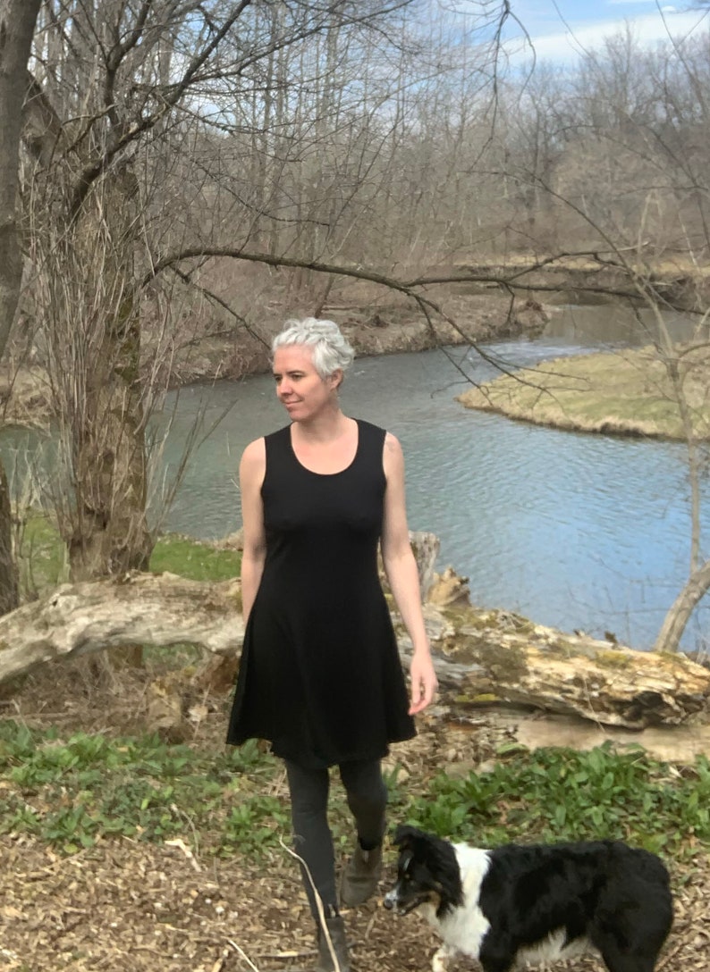 May include: A sleeveless black dress with a scoop neckline and a flared skirt that falls above the knee. The dress is worn with dark gray leggings and brown boots. A black and white dog is visible in the lower foreground of the frame.