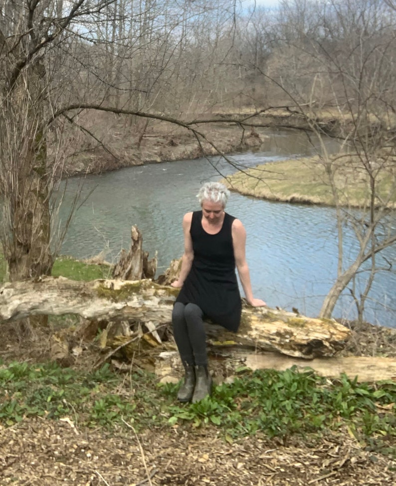 May include: A black sleeveless tank dress worn with dark gray leggings and olive green ankle boots. The dress features a scoop neckline and a simple, fitted bodice that flares slightly at the hem, providing a clean and functional silhouette.
