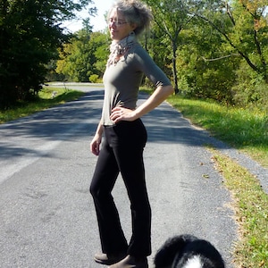 May include: A person wearing a light olive green long-sleeve shirt, black pants, and brown shoes stands on a paved road. A black and white dog is lying down on the road. The background features trees and a blue sky.