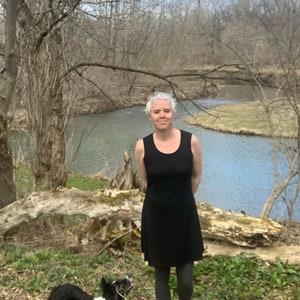 May include: A black sleeveless dress with a scoop neckline and a flared skirt, worn with dark gray leggings and dark boots. A black and white dog with a leash stands nearby, positioned in front of a river and bare trees in a natural outdoor setting.