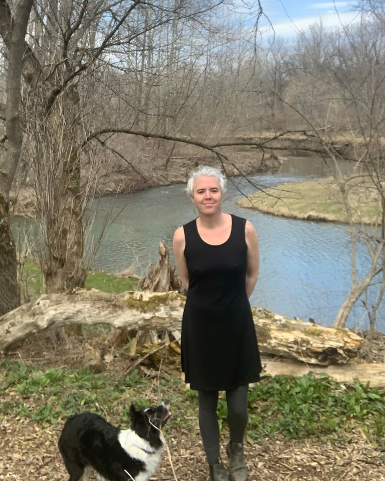 May include: A black sleeveless dress with a scoop neckline and a flared skirt, worn with dark gray leggings and dark boots. A black and white dog with a leash stands nearby, positioned in front of a river and bare trees in a natural outdoor setting.