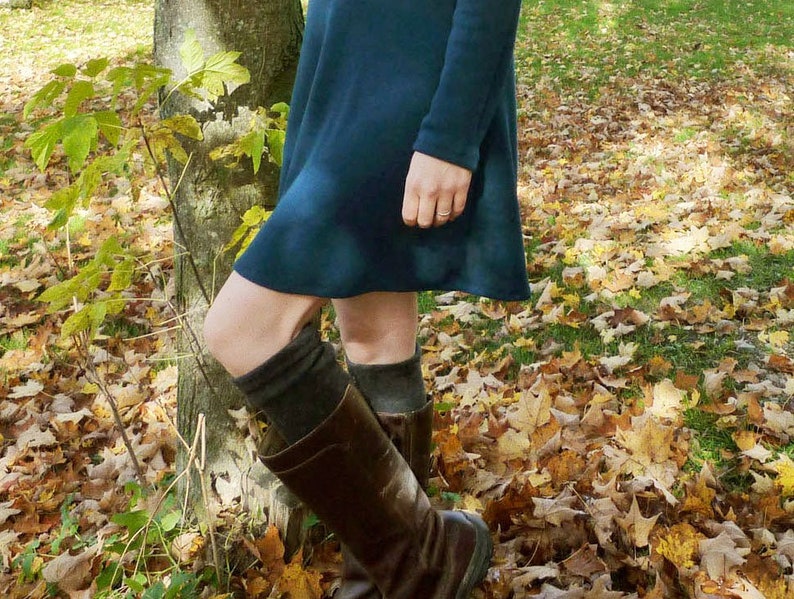K&ouml;nnte beinhalten: Ein dunkelblaugr&uuml;nes Langarmkleid mit ausgestelltem Saum, kombiniert mit dunkelgrauen Strick-Overknee-Socken und braunen Lederstiefeln. Das Outfit zeichnet sich durch ein schlichtes, einfarbiges Design mit glatter Textur am Kleid und gerippten Strickdetails an den Socken aus.