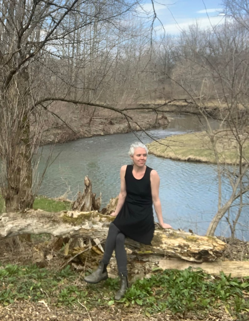 May include: A black sleeveless tank dress worn with dark gray leggings and olive green ankle boots. The dress features a simple scoop neckline and a relaxed, straight-cut silhouette that falls above the knees, providing a clean and functional look.
