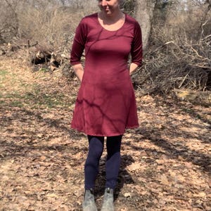 May include: A knee-length, deep burgundy dress with three-quarter length sleeves and a rounded neckline. The dress is paired with dark navy blue leggings and grey ankle boots. The garment features a simple, fitted bodice and a flared skirt.