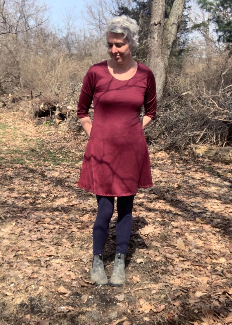 May include: A knee-length, deep burgundy dress with three-quarter length sleeves and a rounded neckline. The dress is paired with dark navy blue leggings and grey ankle boots. The garment features a simple, fitted bodice and a flared skirt.