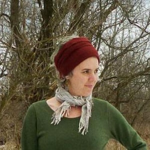 Puede incluir: Una mujer con un turbante de punto burdeos, una camisa verde de manga larga y una bufanda gris con flecos. El fondo presenta árboles desnudos y un indicio de nieve, lo que sugiere un entorno frío al aire libre.