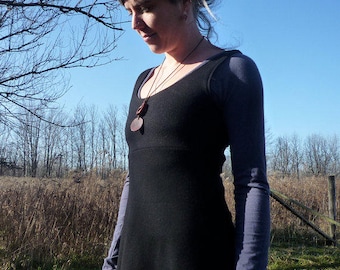 May include: A black sleeveless dress with a scoop neckline worn over a long-sleeved slate blue shirt. A brown circular pendant hangs from a thin cord around the neck. The dress features a fitted bodice that transitions into a flared skirt silhouette.