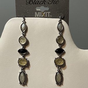 Könnte beinhalten: Lange Ohrhänger mit silberfarbenem Metallfinish. Jeder Ohrring hat eine Reihe von ovalen und runden Steinen in Weiß, Schwarz und Gelb. Die Ohrringe werden auf einer weißen Oberfläche mit einem "Black Tie"-Etikett präsentiert.
