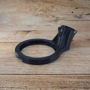 Könnte beinhalten: Ein schwarzes 3D-gedrucktes Objekt mit einem kreisförmigen Ring und einer vertikalen Komponente. Auf dem Ring ist der Text "www.3D Printsolutions" aufgedruckt. Das Objekt befindet sich auf einer Holzoberfläche.